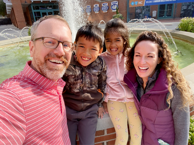 Two smiling adults and two laughing children pose close together for a selfie in front of an outdoor fountain.