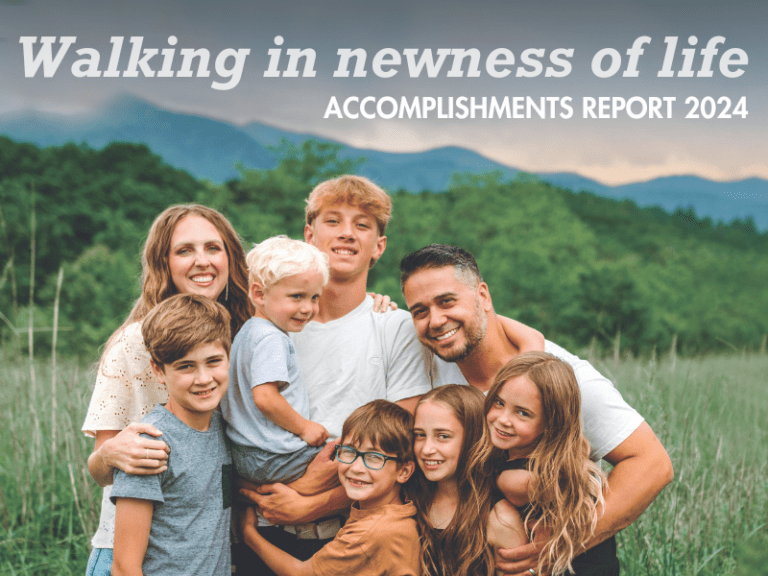Smiling family of eight hugging in a grassy field with mountains behind, on the "Walking in newness of life" 2024 Accomplishments Report cover
