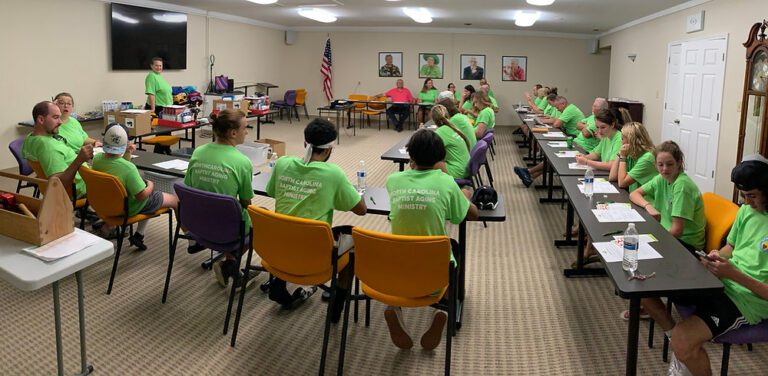 Volunteers in green "North Carolina Baptist Aging Ministry" shirts sit at long tables in a training session while a leader stands at the front.