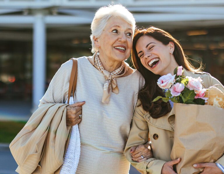 An older woman and a younger woman laugh together arm in arm, carrying groceries and pink roses outdoors.