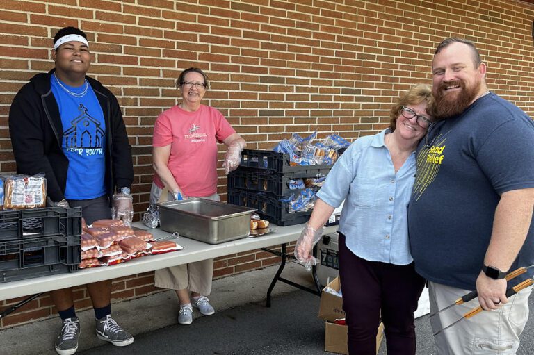 Four volunteers smile beside a table of hot dogs and buns at an outdoor community meal, one wearing a "Send Relief" shirt.