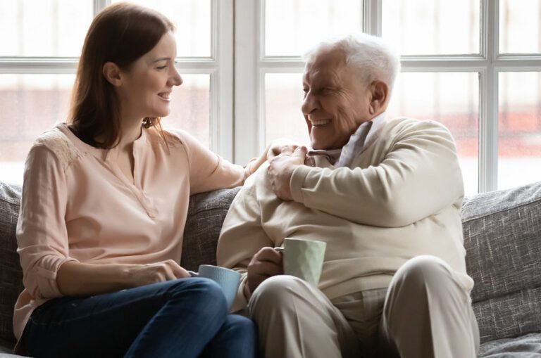 A smiling young woman and an older man laugh together on a couch while holding mugs near a bright window.