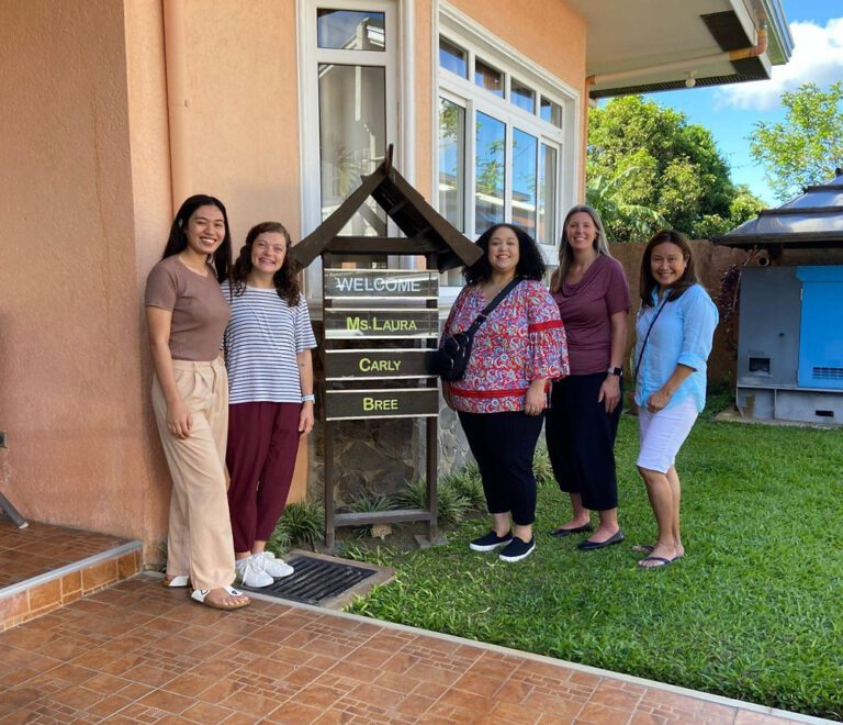 Five smiling women pose beside a wooden welcome sign outside a peach building with a green lawn and tropical trees.