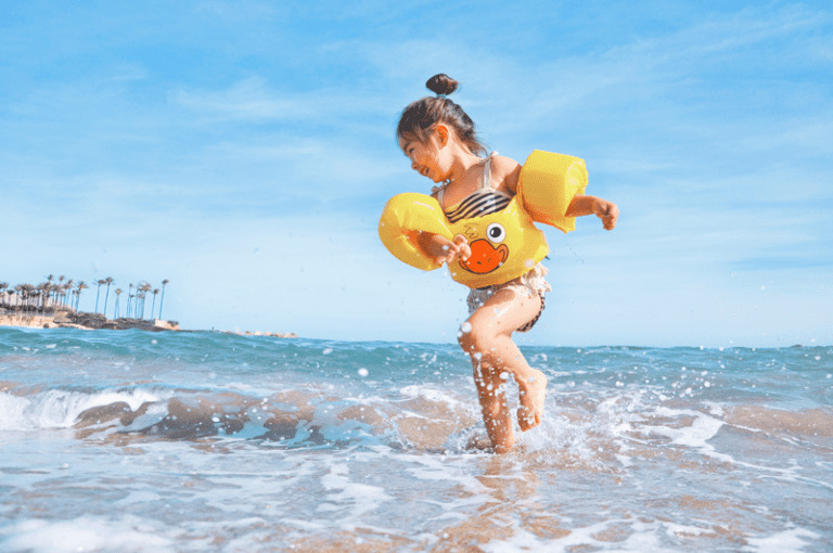 Smiling young girl in a yellow duck swim vest splashes through shallow ocean waves on a sunny beach day.