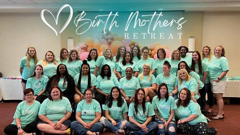 Women in matching mint "Loved" shirts pose together at the Birth Mothers Retreat, with balloons behind them.