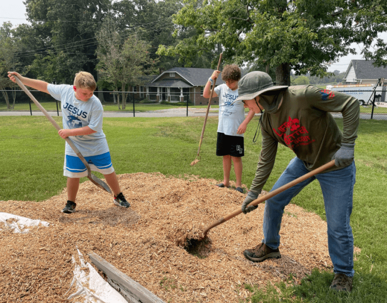 Two boys in "Jesus" shirts and an adult in a hat spread wood mulch with rakes and shovels on a lawn.