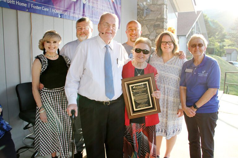 Seven adults smile together outdoors as an older woman in sunglasses holds a framed commemorative plaque.