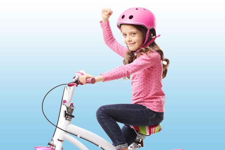 Smiling girl in a pink helmet and polka-dot top raises her fist in triumph while riding a white bicycle.