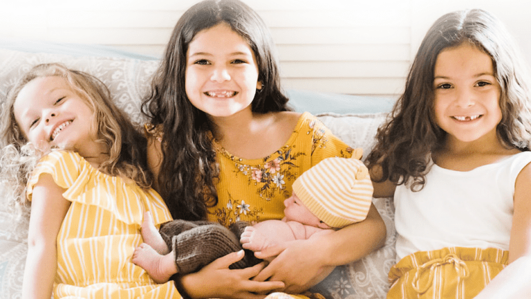 Three smiling girls in yellow and white sit on a couch, the middle girl holding a newborn in a striped hat.