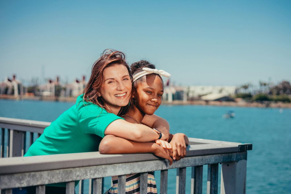 A mother and daughter share a joyful moment by the water on a sunny day.
