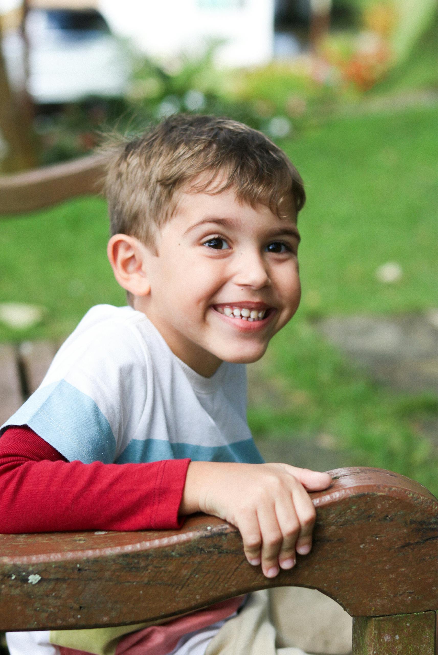 Playful young boy sitting on a wooden bench outdoors, smiling brightly.