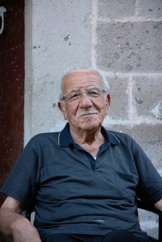 Portrait of a senior man with eyeglasses sitting against a brick wall.