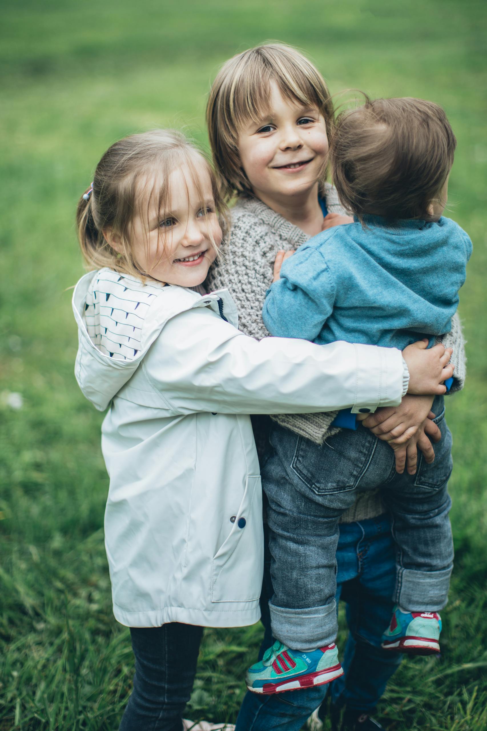 Three cheerful siblings embracing in a green field on a sunny day, showcasing their joyful bond.