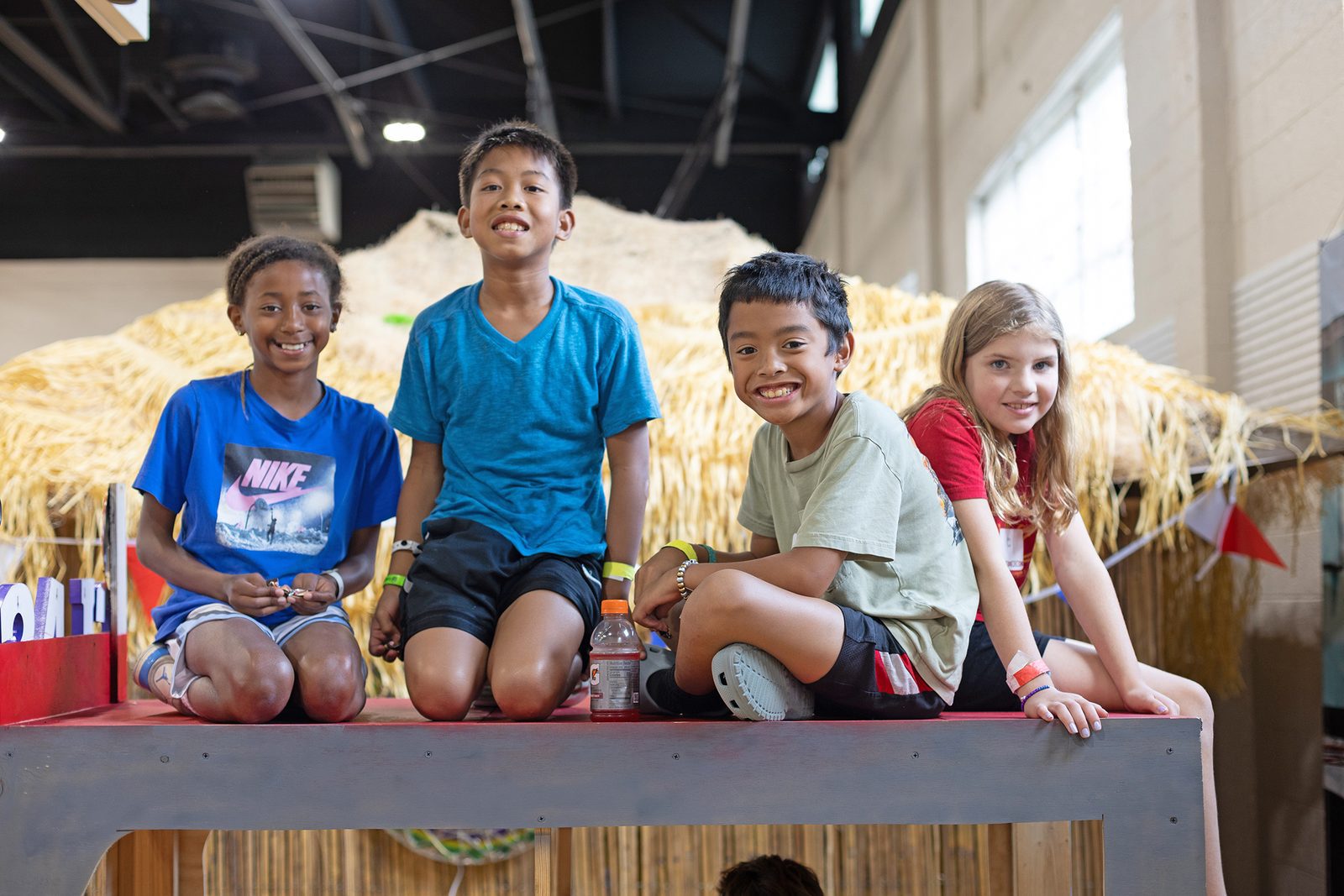 A group of children sitting together outdoors on a wooden structure, smiling at the camera.