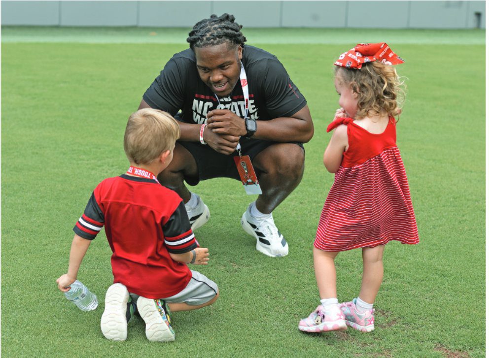 NC State inside linebacker AJ Richardson kneels on the field talking with two young children from Baptist Children’s Homes