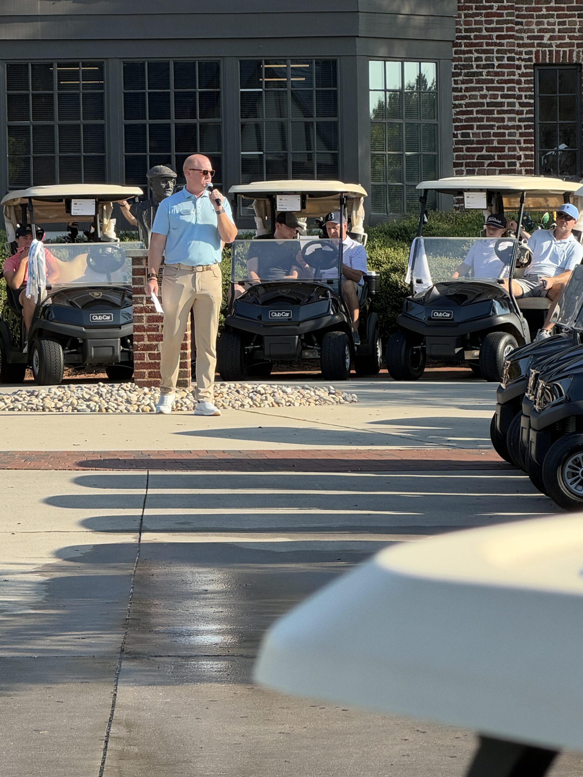 Man in a light blue polo speaks into a microphone before rows of golfers seated in Club Car golf carts at a tournament.
