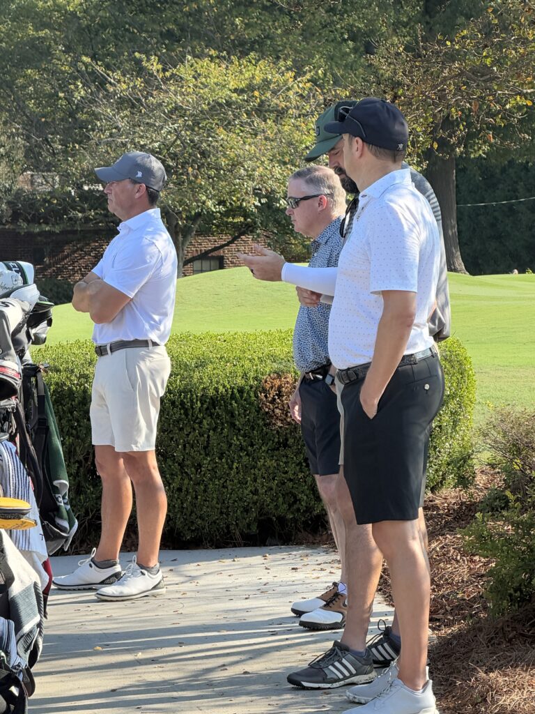 Four men in golf attire stand together on a cart path beside a green hedge, with golf bags at left and a fairway behind.
