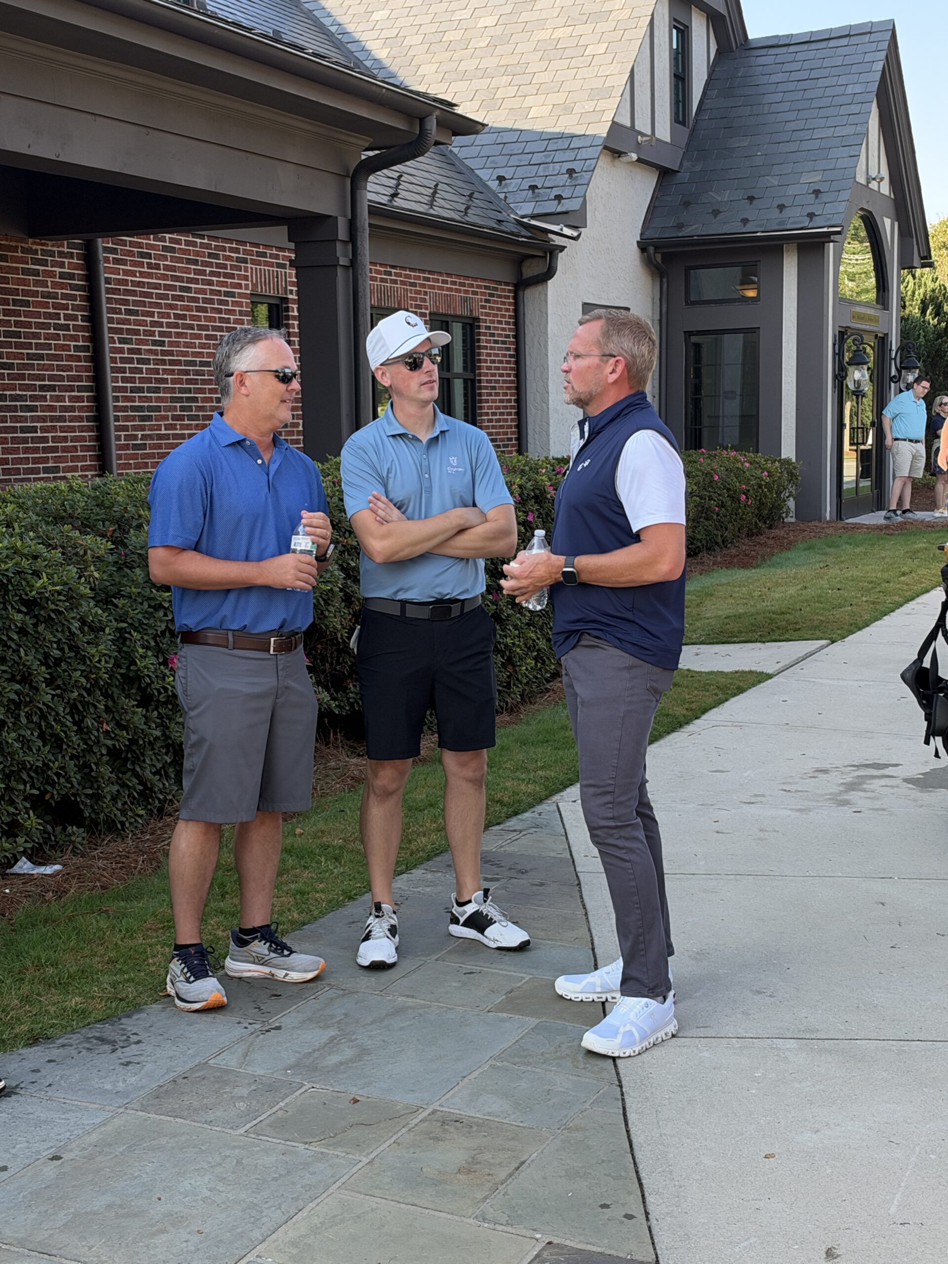 Three men in golf attire stand talking on a sidewalk outside a brick clubhouse, holding water bottles on a sunny day.
