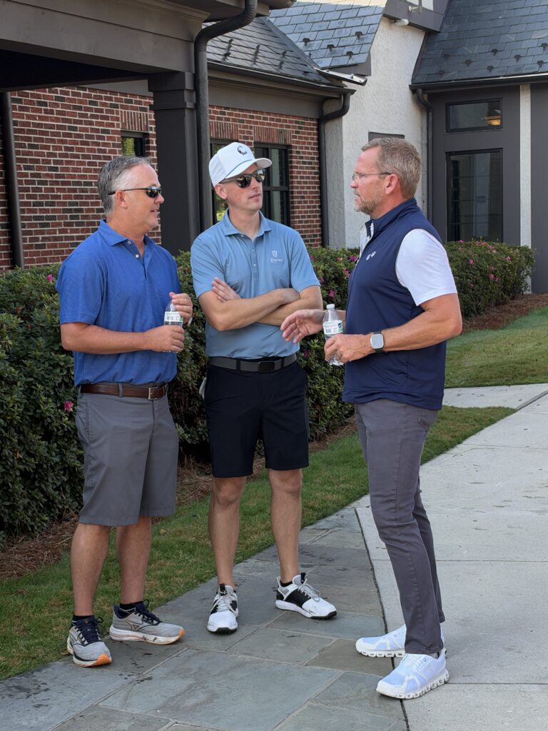 Three men in golf attire stand talking outdoors on a stone walkway, holding water bottles near a brick building.