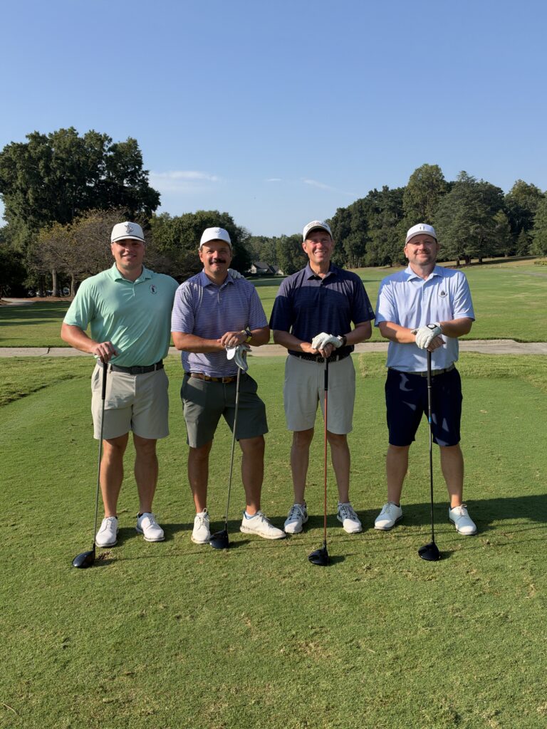 Four men in golf attire and caps stand smiling on a sunny golf course tee box, each holding a golf club.
