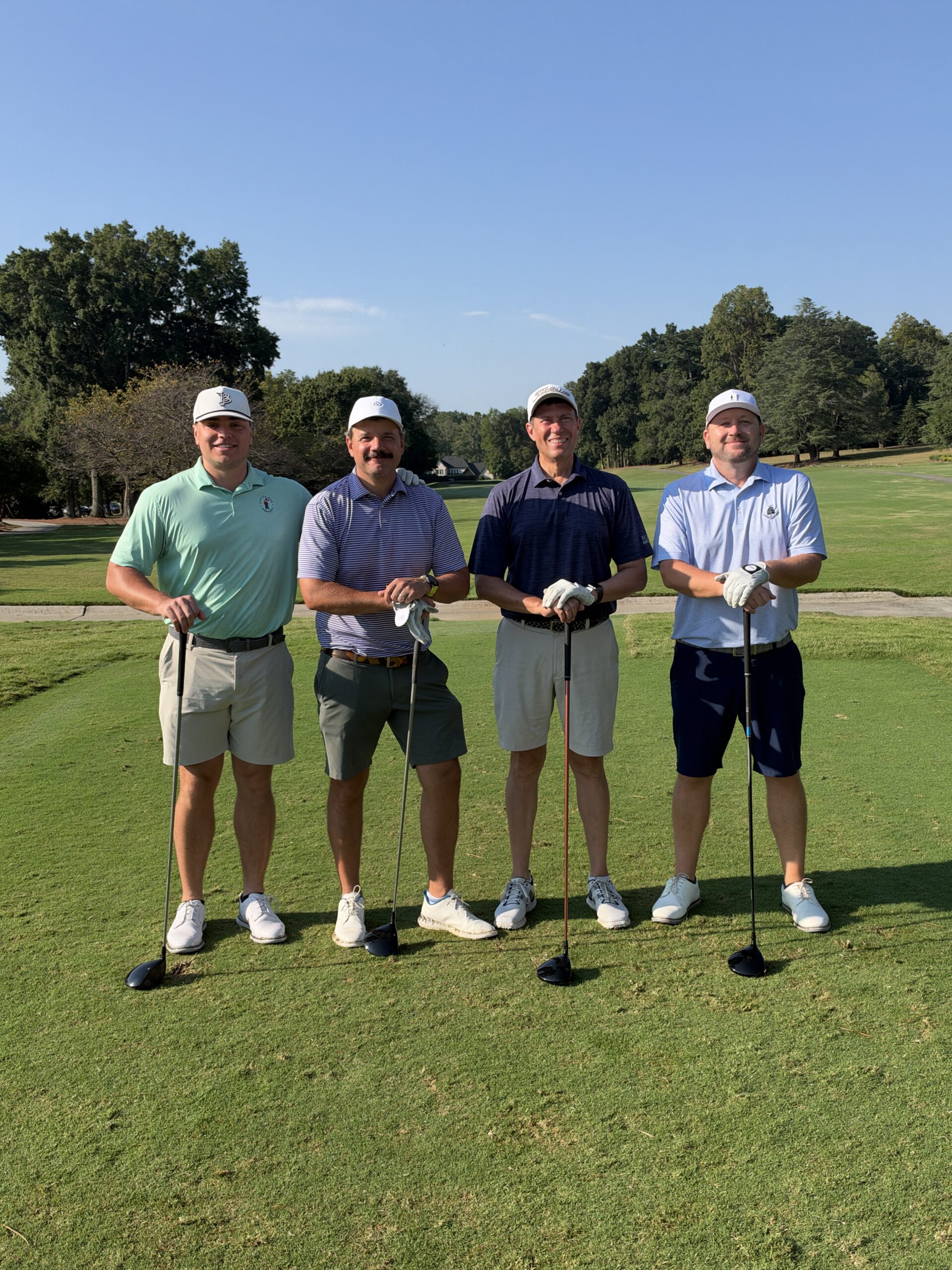 Four men in golf attire and caps stand smiling on a sunny golf course tee box, each holding a golf club.