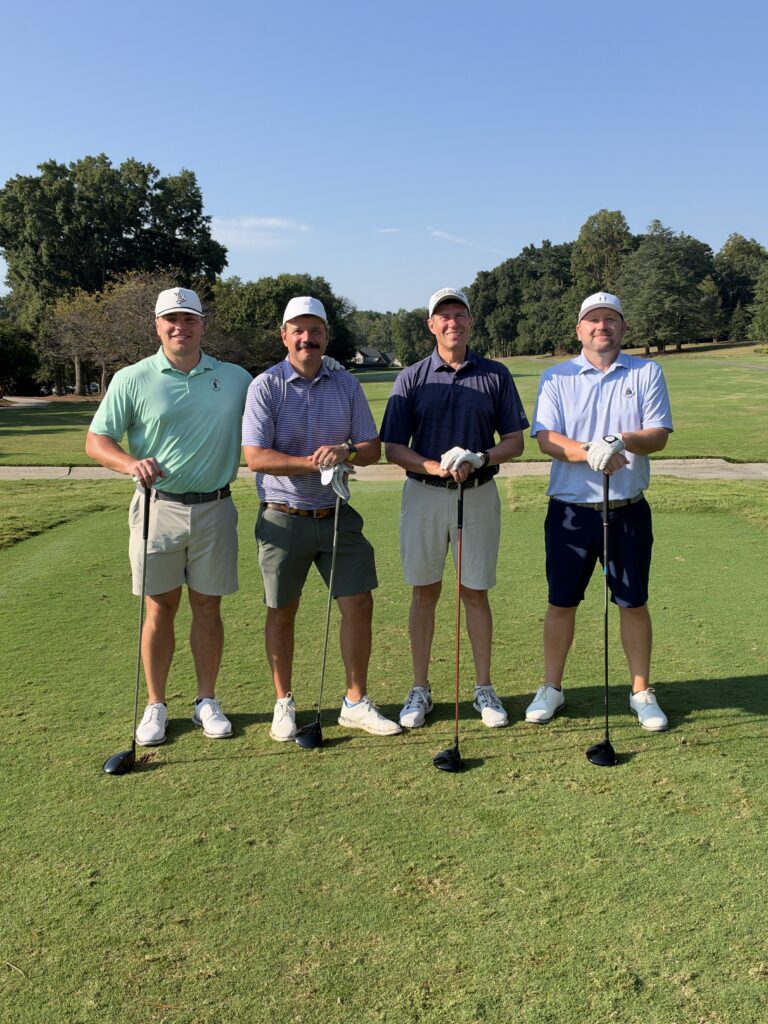 Four men in golf attire and white caps pose with their clubs on a sunny golf course tee box.