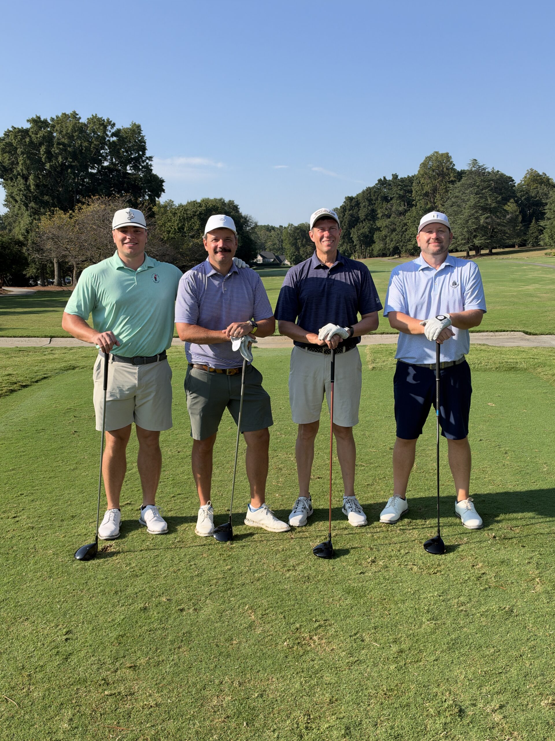 Four men in golf attire and white caps pose with their clubs on a sunny golf course tee box.
