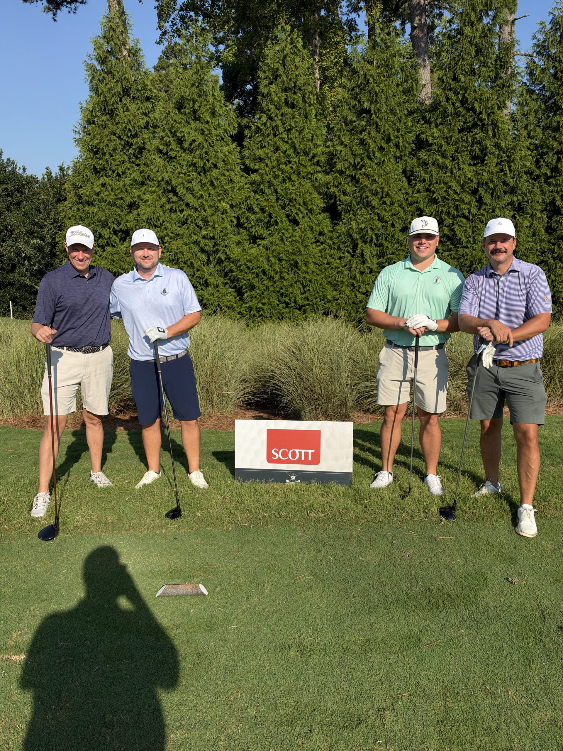 Four men in golf attire pose with clubs on a tee box beside a red and white "Scott" sponsor sign.