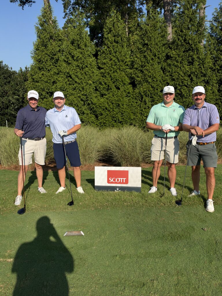 Four golfers pose with clubs on a tee box behind a red and white "Scott" sign on a sunny day.