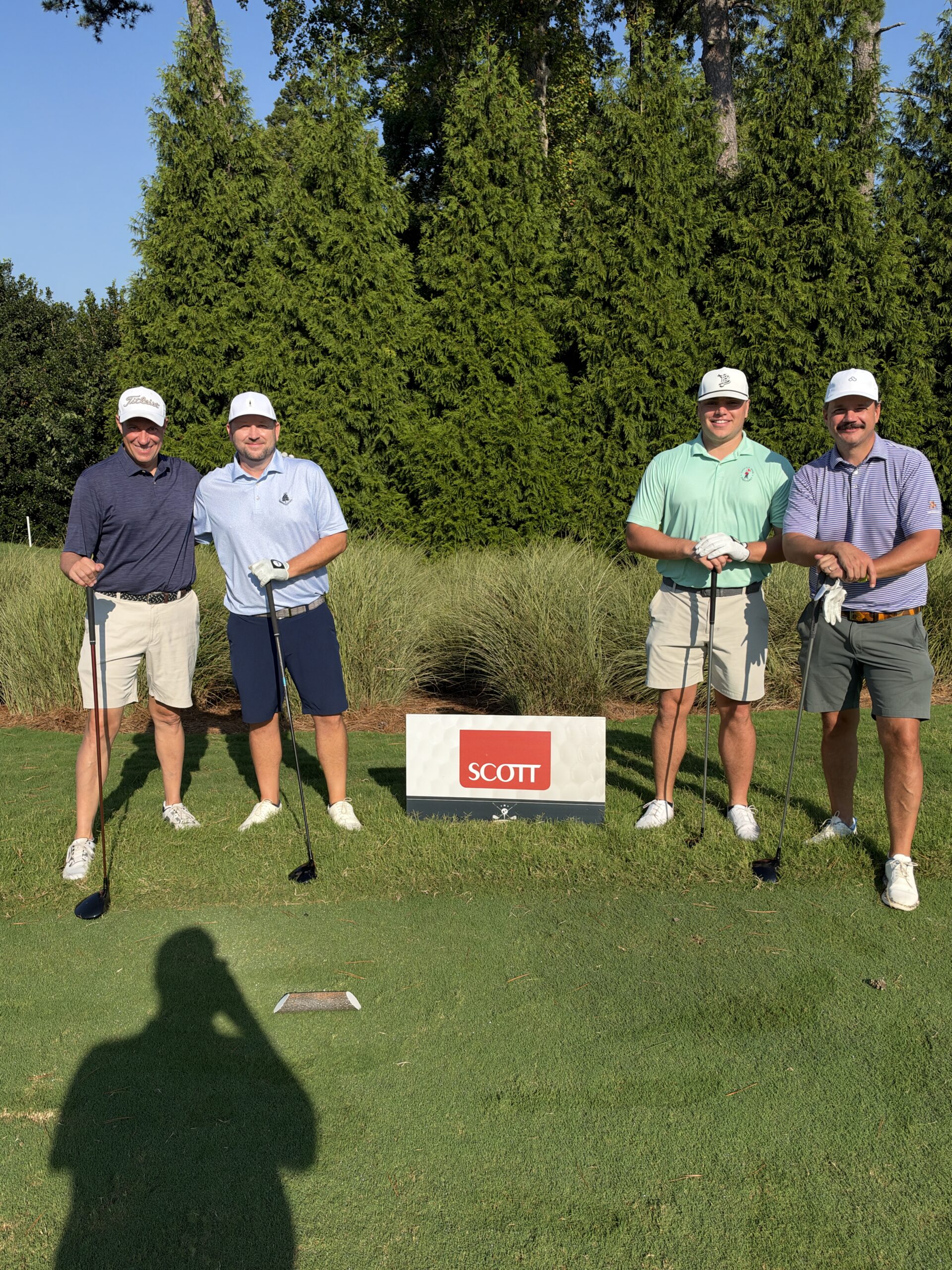 Four golfers pose with clubs on a tee box behind a red and white "Scott" sign on a sunny day.