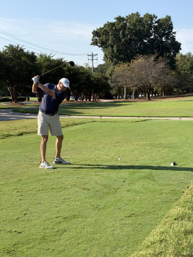 Golfer in a navy polo and white cap finishes a backswing at a tee box on a sunny golf course lined with trees.