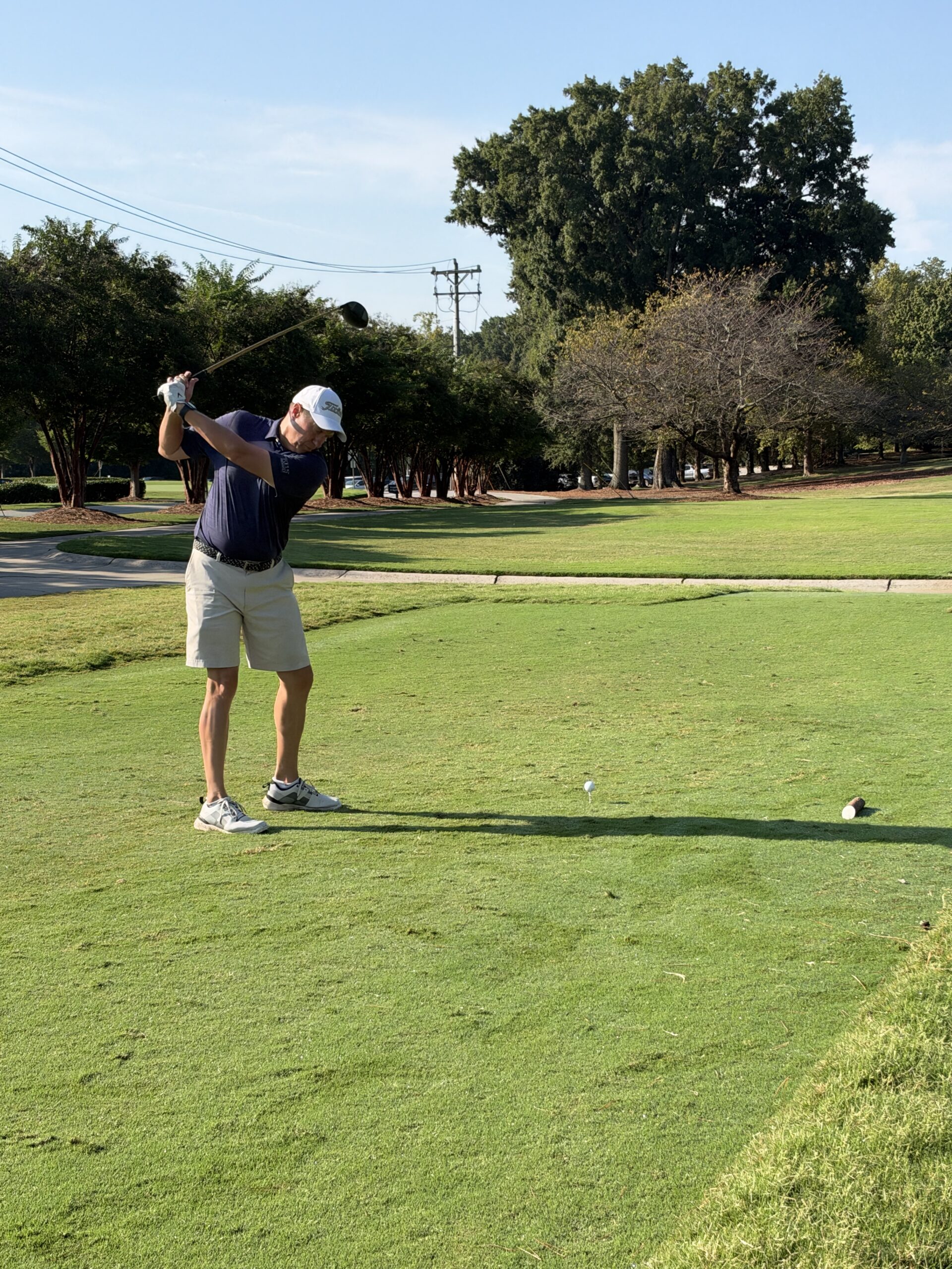 Golfer in a navy polo and white cap finishes a backswing at a tee box on a sunny golf course lined with trees.