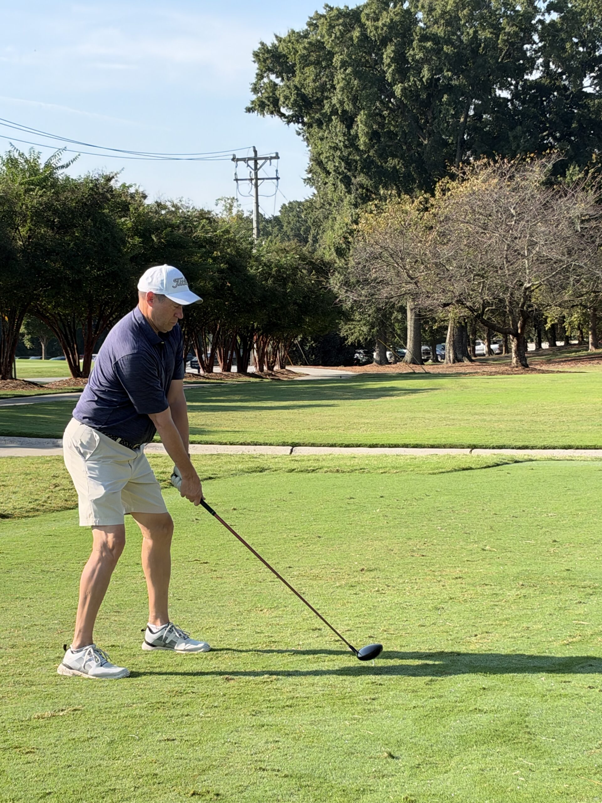 Man in a white cap, navy polo and khaki shorts addresses the ball with a driver on a sunny golf course tee.