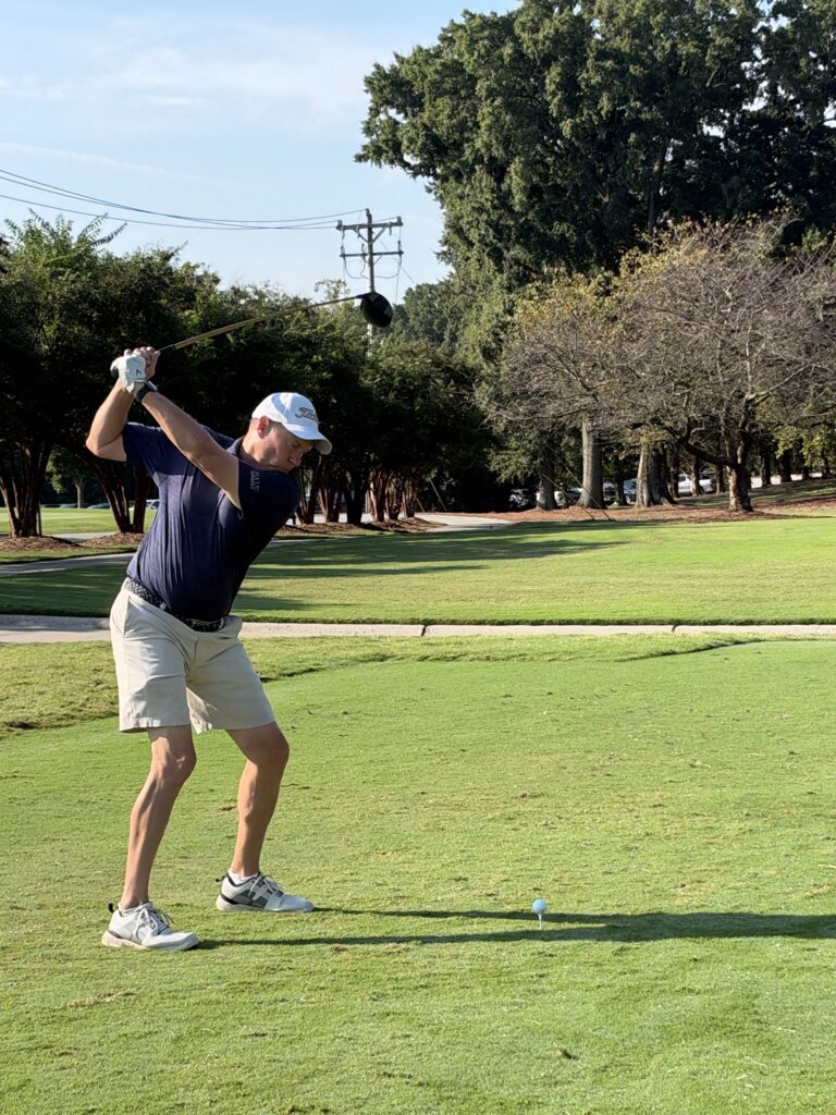 Golfer in a navy polo, khaki shorts and white cap at the top of a backswing on a sunny tee box, with a ball on the tee.