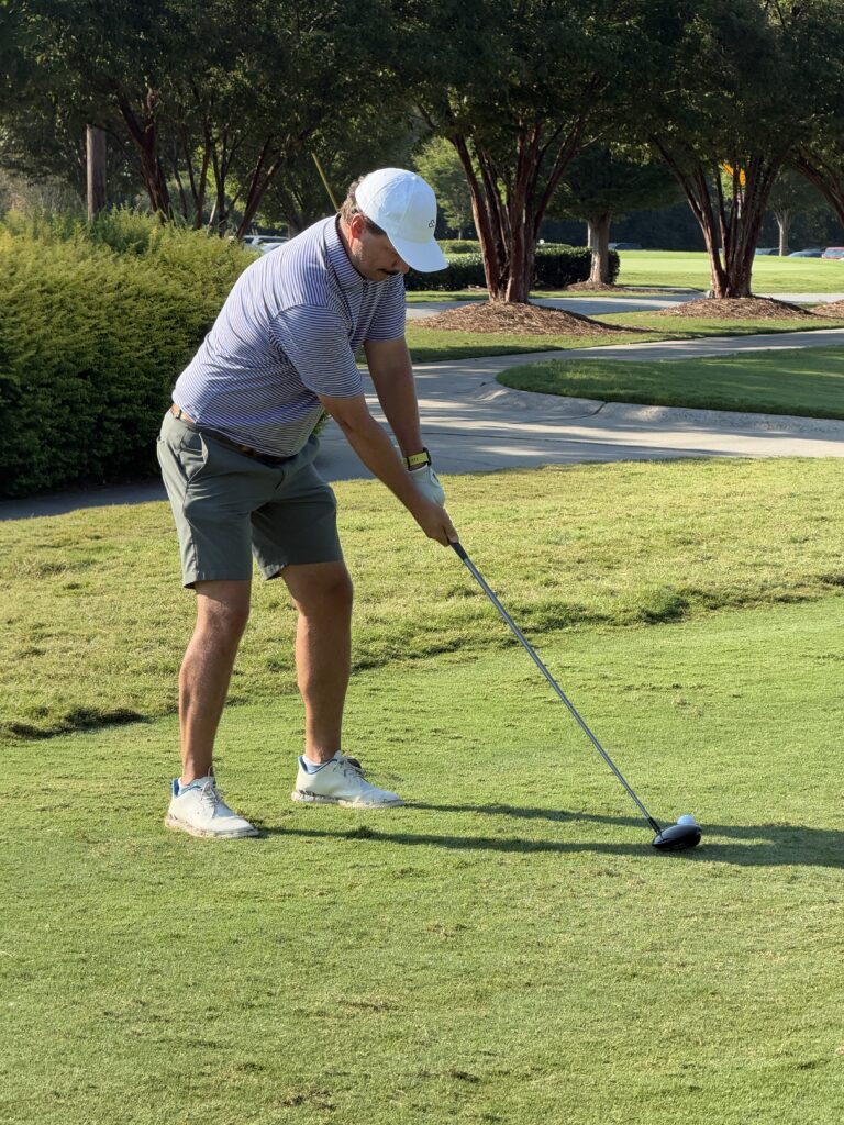 Golfer in a white cap and striped polo lines up a tee shot with a driver on a sunny, tree-lined golf course.