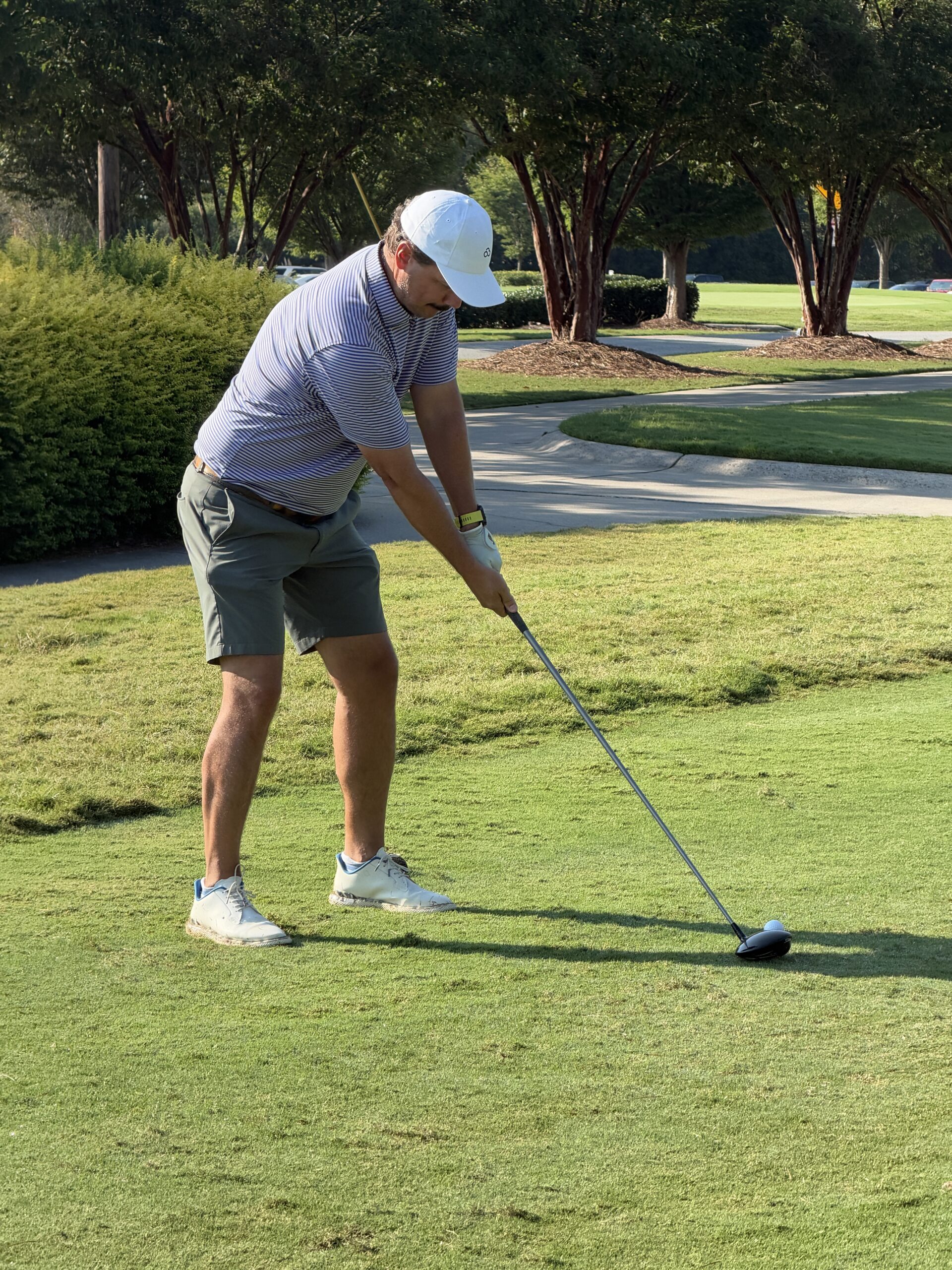 Golfer in a white cap and striped polo lines up a tee shot with a driver on a sunny, tree-lined golf course.