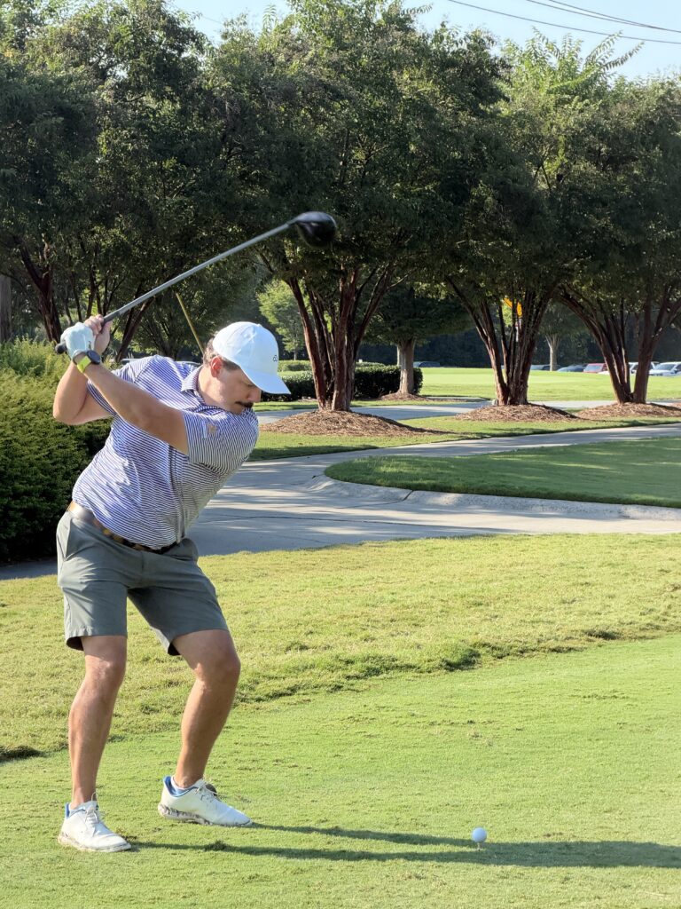 Golfer in a white cap and striped polo at the top of a backswing on a tee, with a golf ball on the grass below.