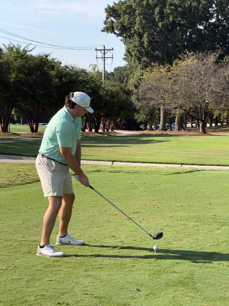 Golfer in a mint polo, khaki shorts and white cap lines up a drive from the tee on a sunny, tree-lined course.