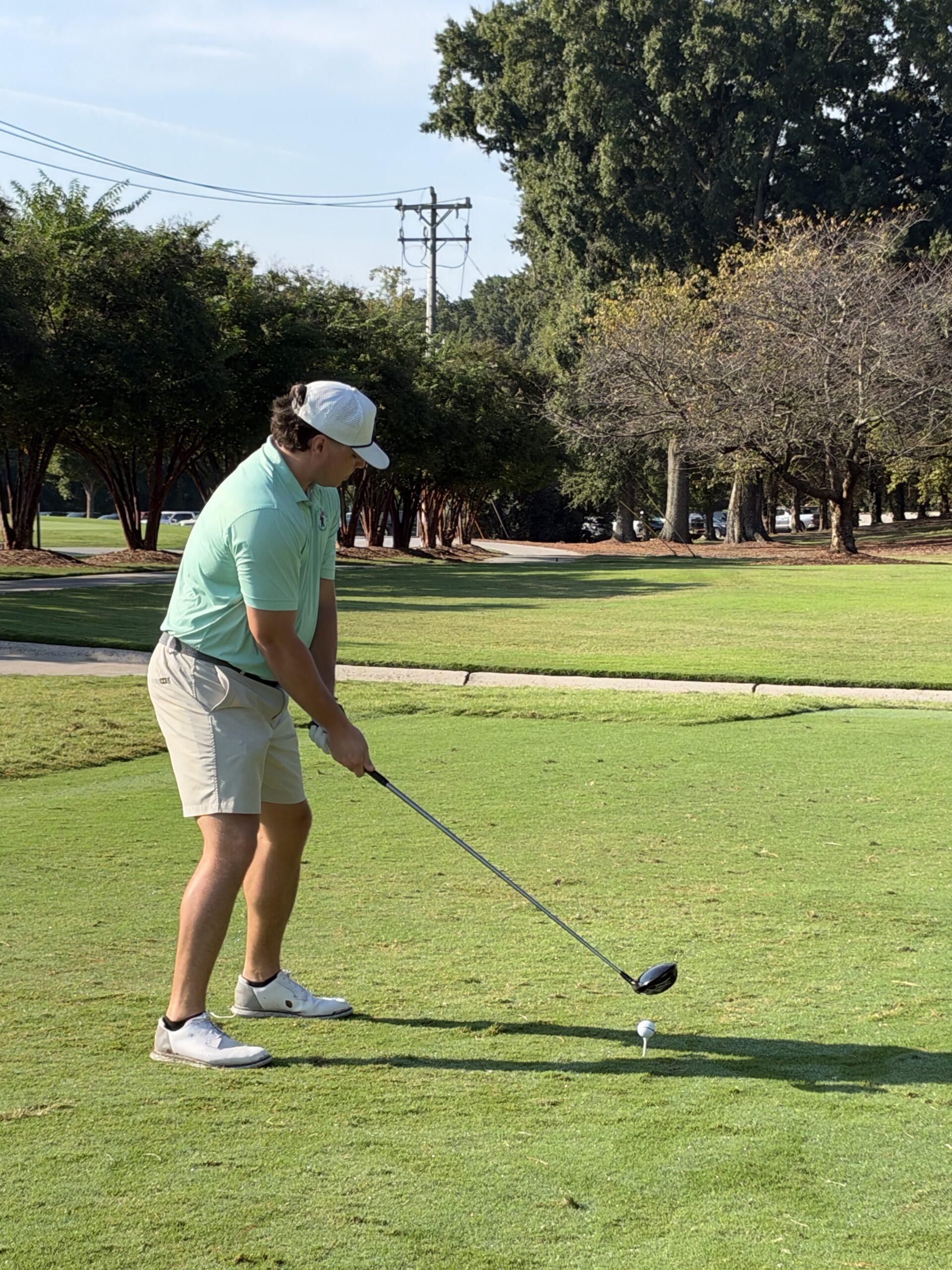 Golfer in a mint polo, khaki shorts and white cap lines up a drive from the tee on a sunny, tree-lined course.