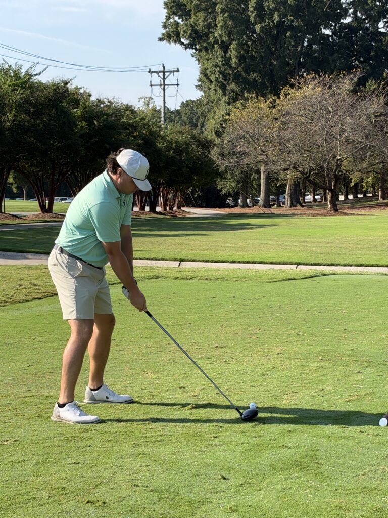 Golfer in a mint polo, khaki shorts and white cap addresses a ball on the tee with a driver on a sunny course.