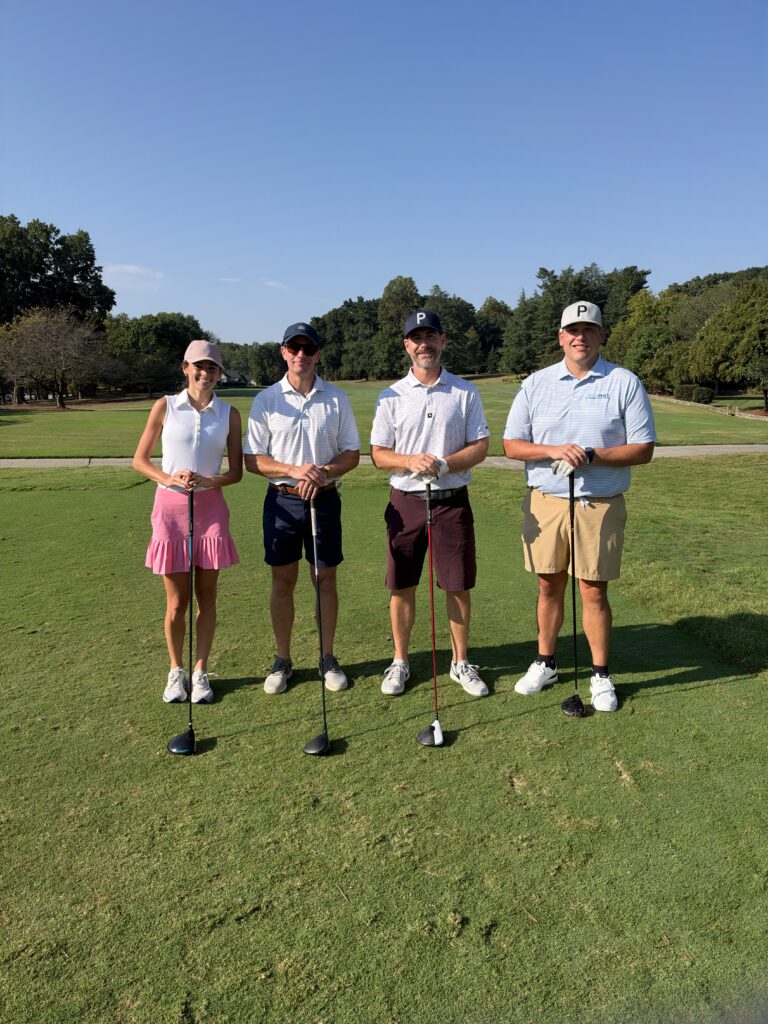 Four golfers, one woman and three men, pose holding clubs on a sunny golf course fairway.