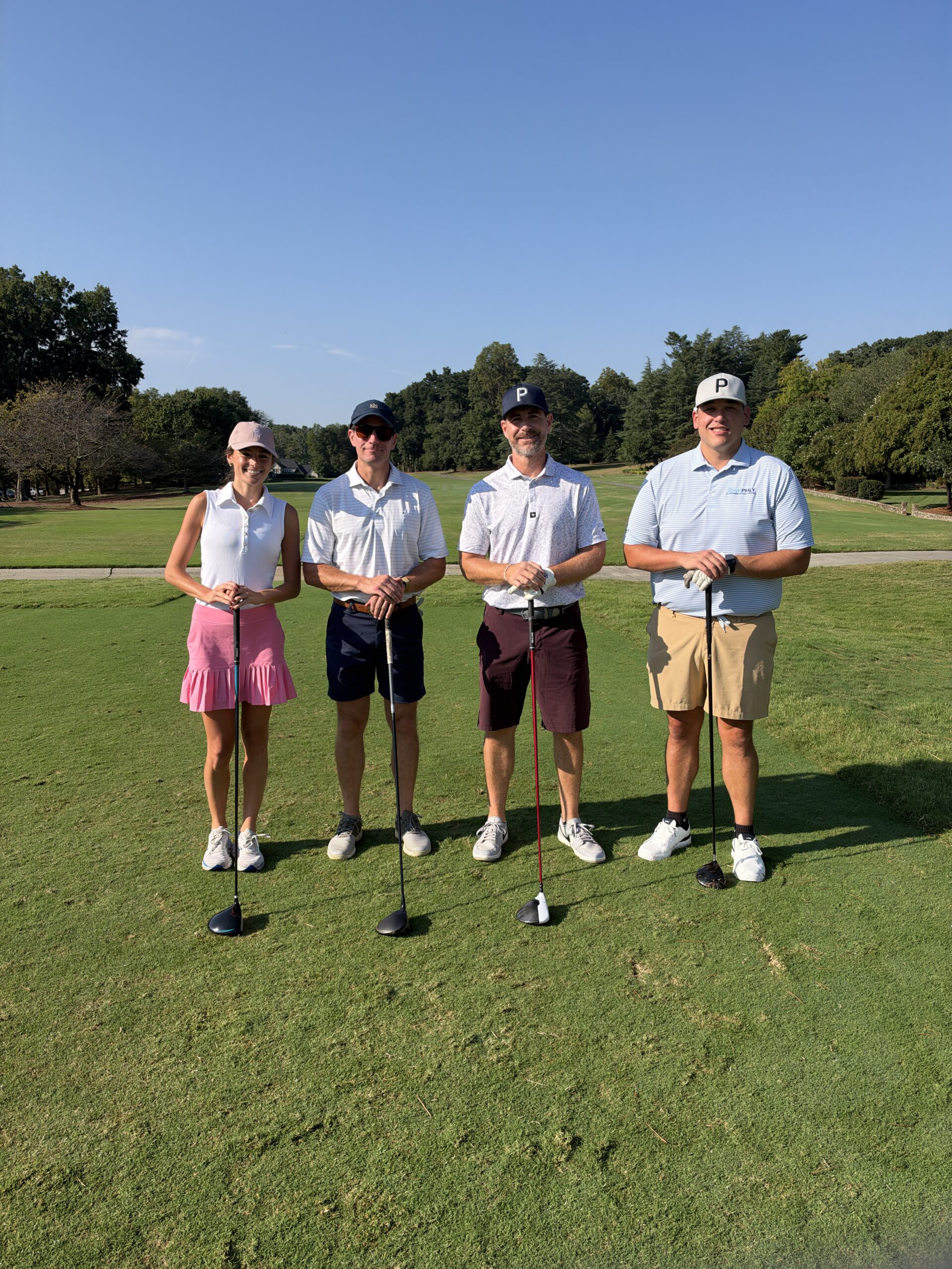 Four golfers, one woman and three men, pose holding clubs on a sunny golf course fairway.
