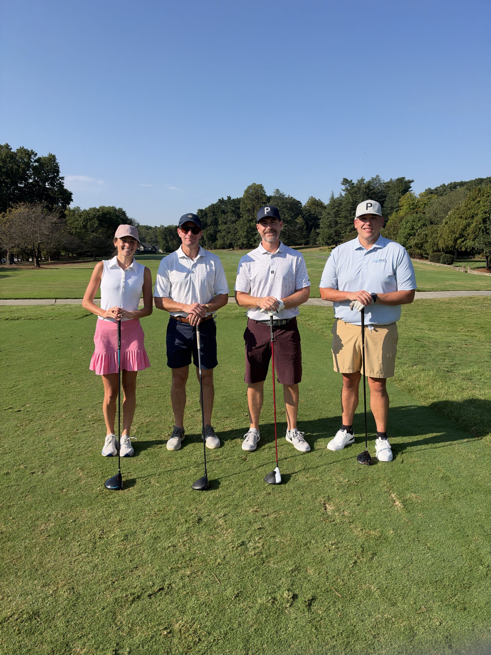 Four golfers, one woman and three men, pose with their clubs on a sunny golf course fairway.