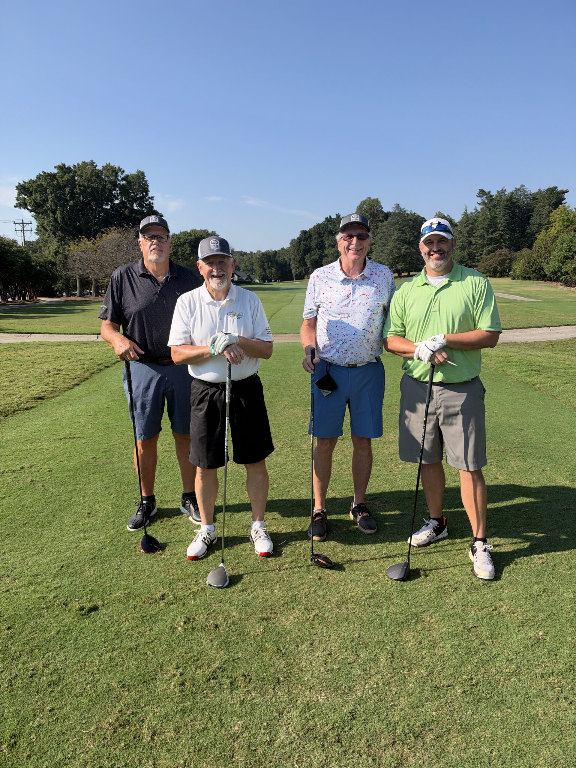 Four smiling men in golf attire stand together on a sunny fairway, each holding a golf club.