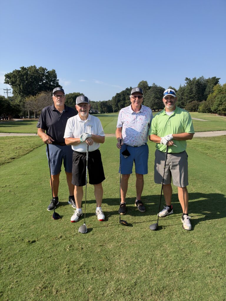 Four men in golf attire pose smiling on a sunny golf course tee box, each holding a club.