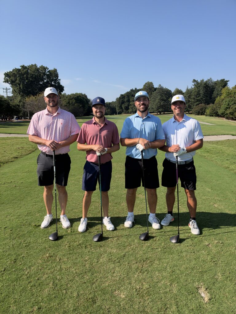 Four men in golf attire stand smiling on a sunny fairway, each leaning on a golf club.