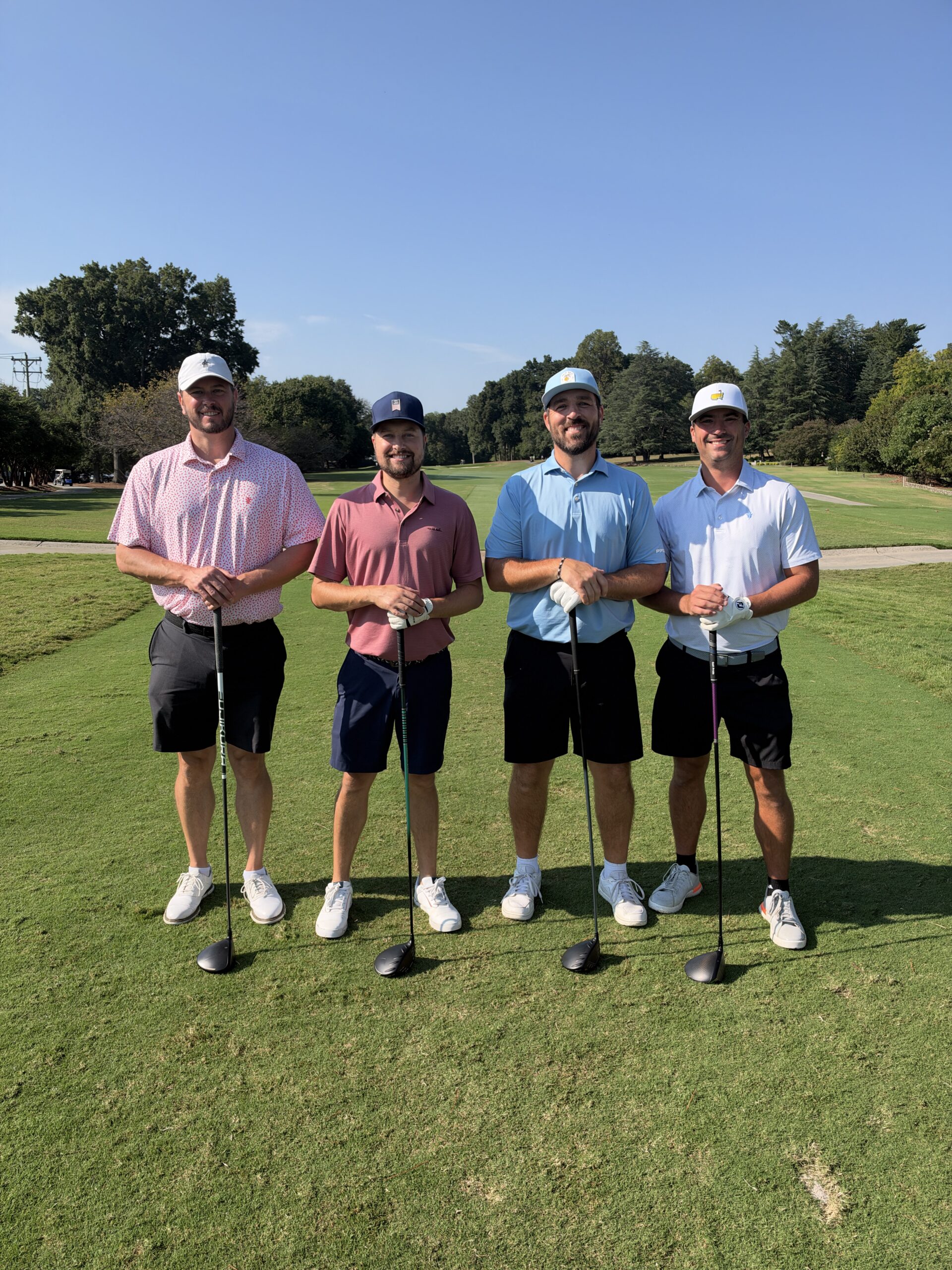 Four men in golf attire stand smiling on a sunny fairway, each leaning on a golf club.