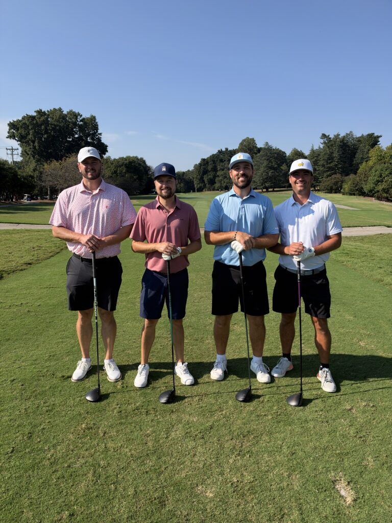 Four men in polos, shorts and caps pose on a sunny golf course fairway, each leaning on a golf club.