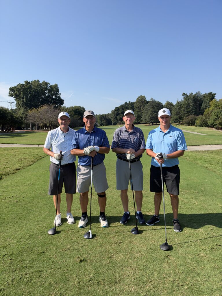 Four men in golf attire stand smiling on a sunny fairway, each leaning on a golf club.