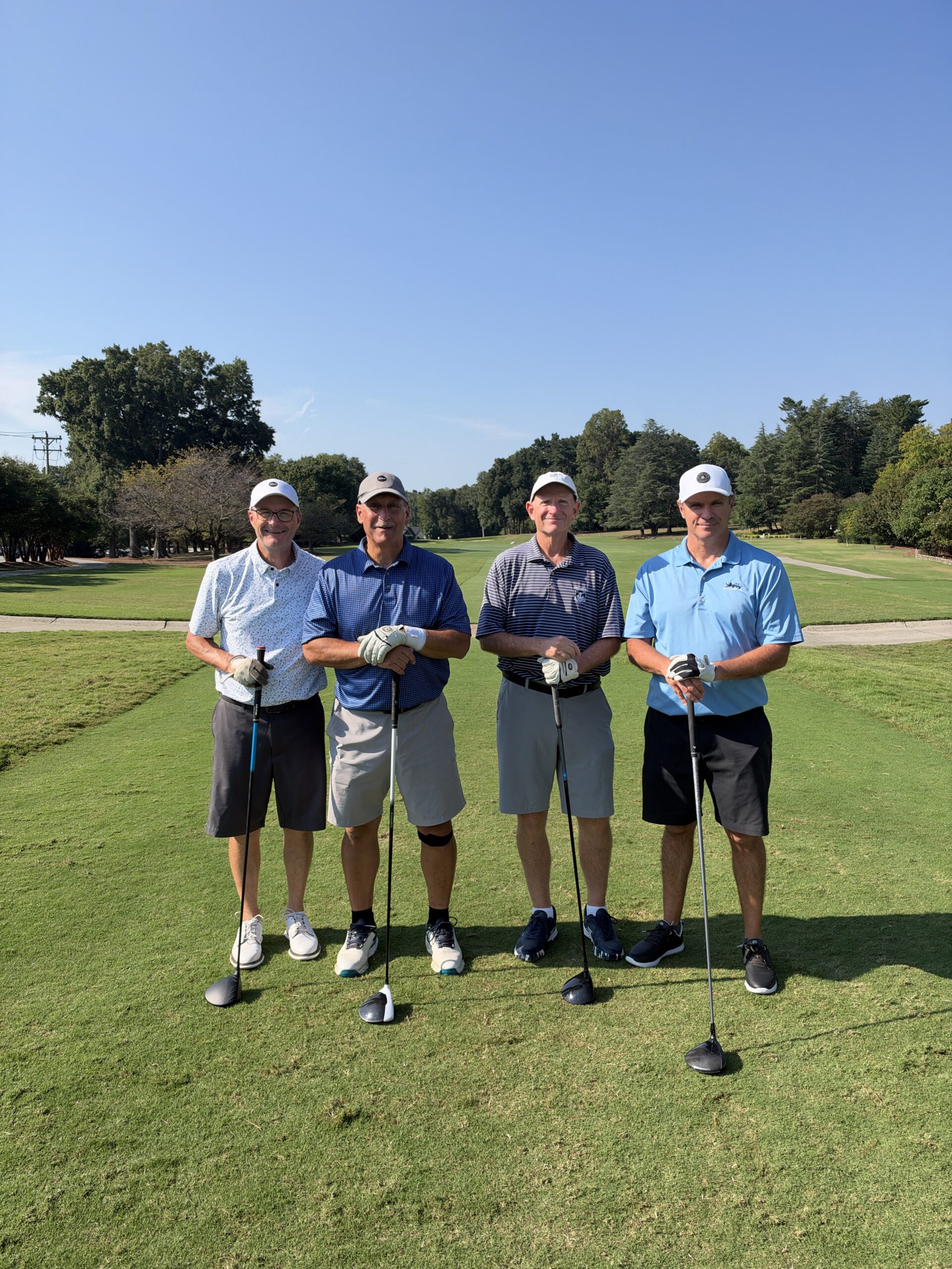 Four men in golf attire stand smiling on a sunny fairway, each leaning on a golf club.